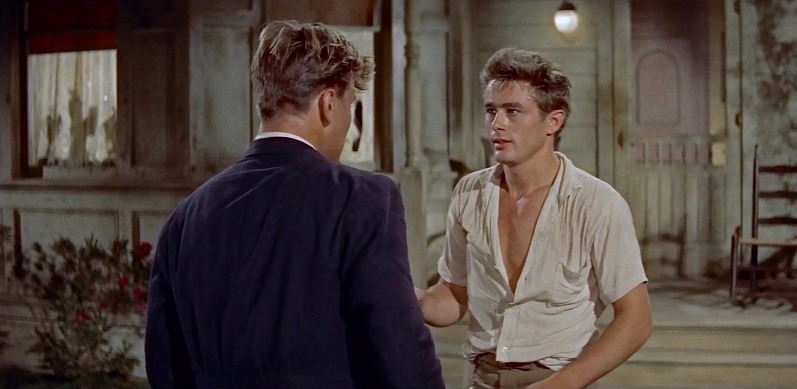 a screenshot from East of Eden showing James Dean with an open white shirt in a suburban outdoor area